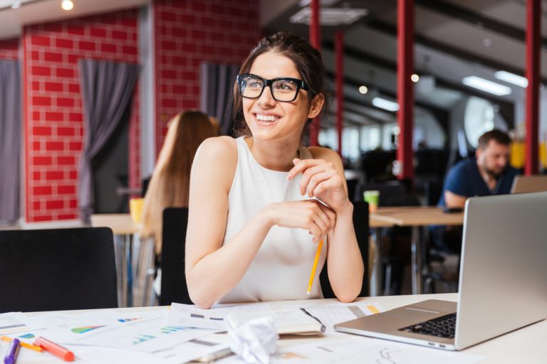 mujer feliz con gafas trabajando con apuntes, ordenador y bolígrafo en un bar