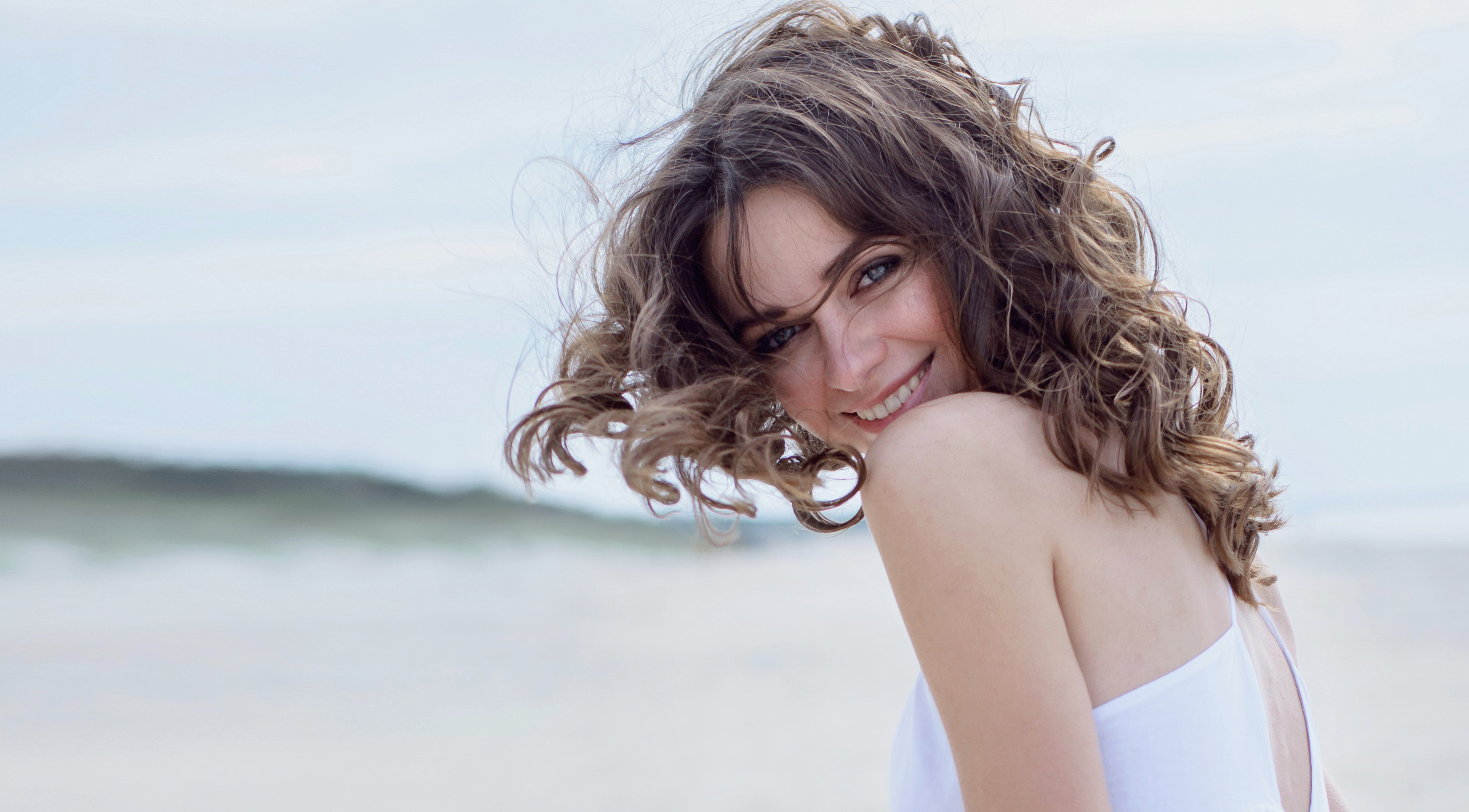 mujer feliz con pelo rizado y de blanco en la playa mirando 