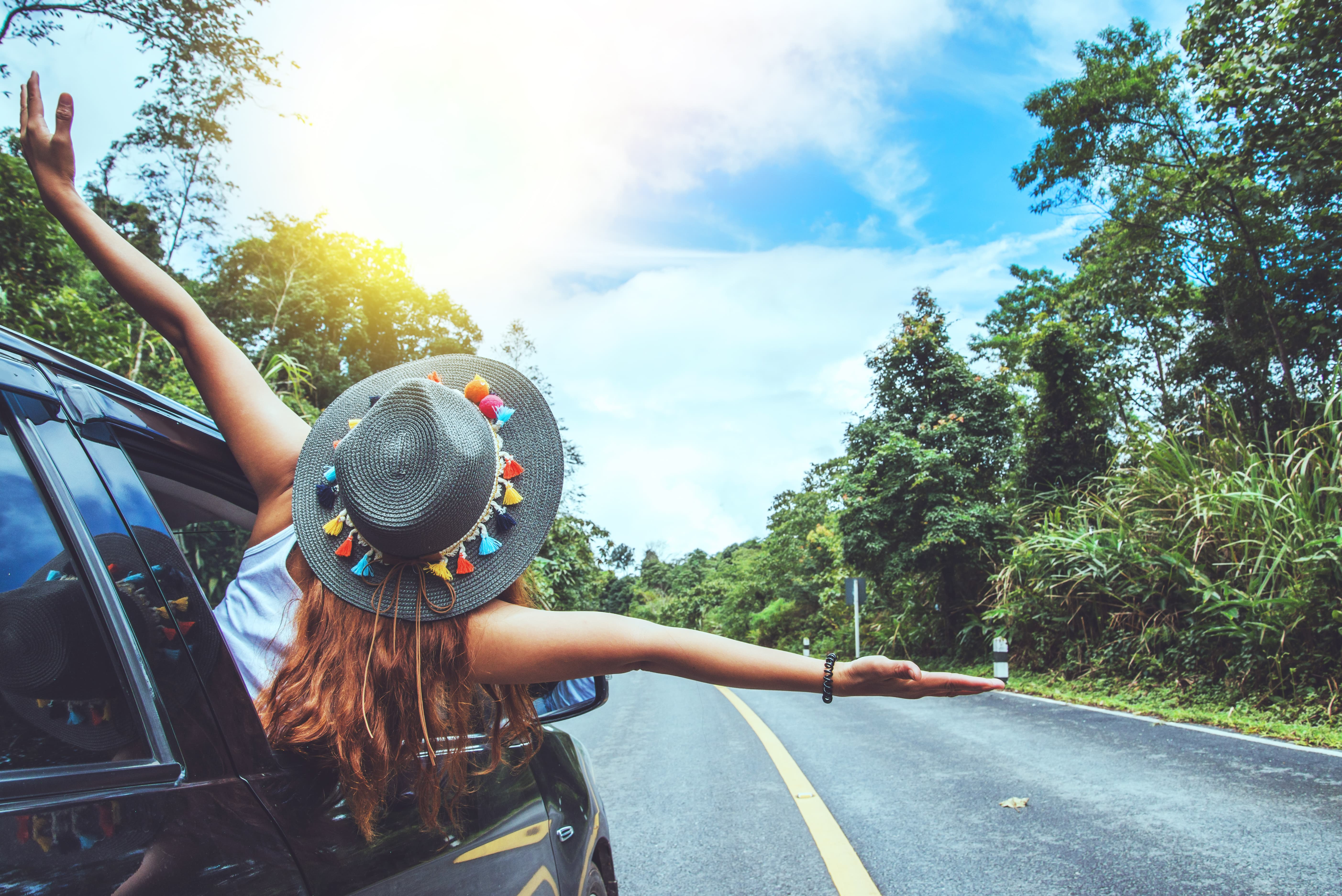 mujer con sombrero sacando el cuerpo por la ventanilla del coche levantando los brazos y mirando al cielo con mucha felicidad en una carretera con mucha naturaleza