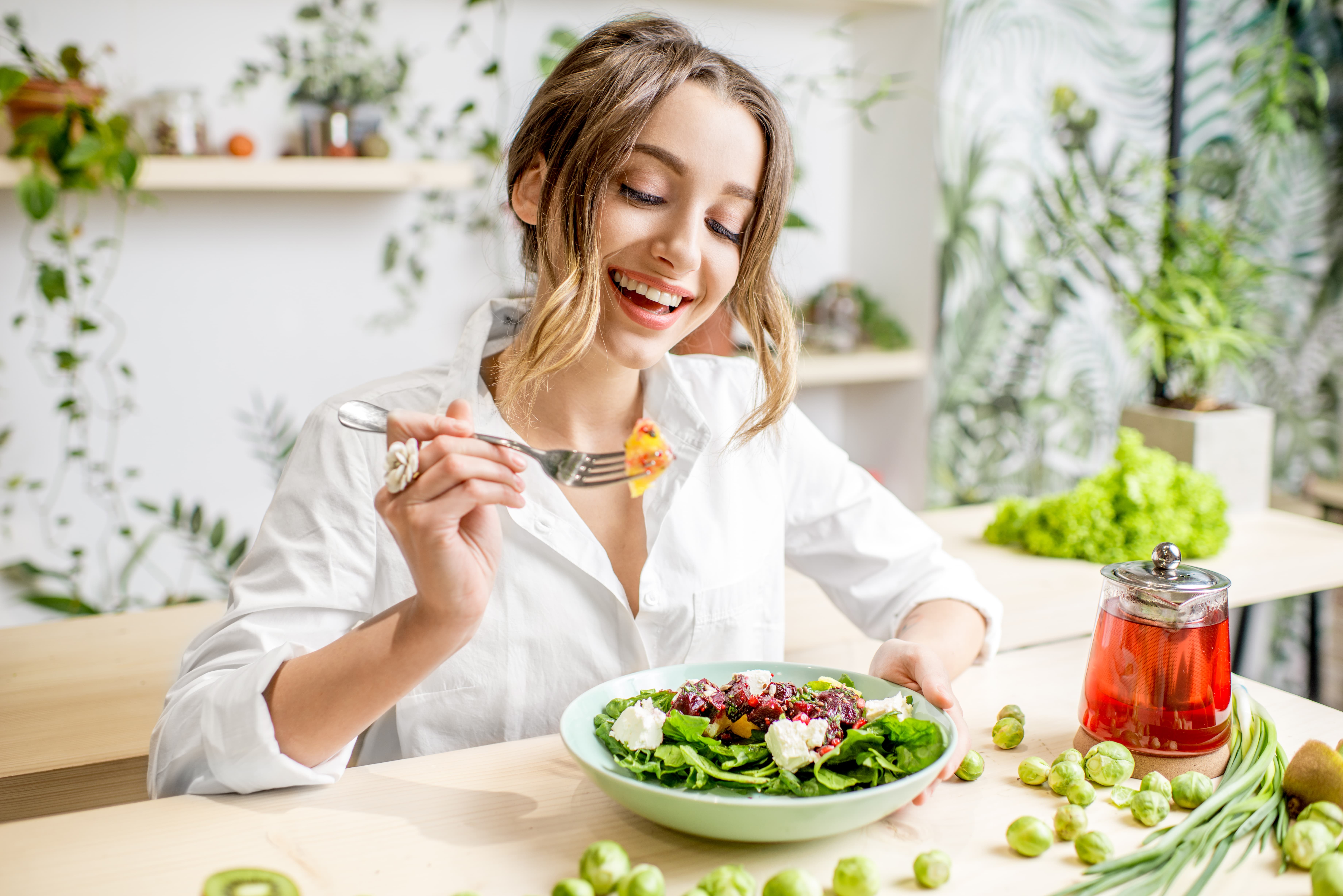 mujer rubia feliz vestida de blanco comiendo una ensalada saludable en un espacio decorado bonito con colores claros y mucha naturaleza